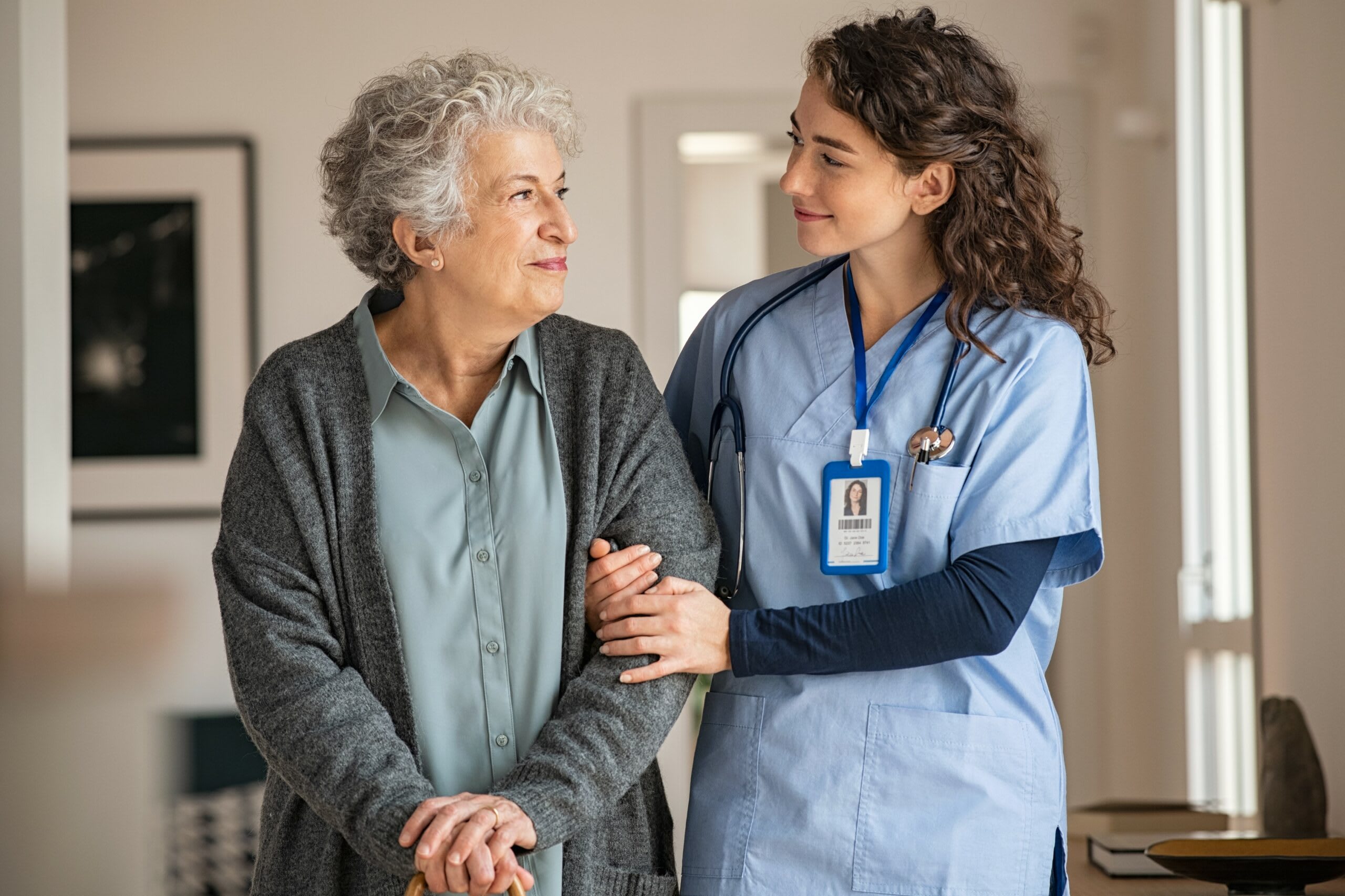 A senior woman and a woman in scrubs walk down a hallway while smiling.